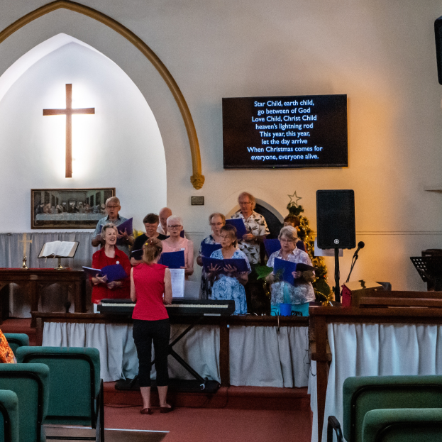 Eine Gruppe von Menschen steht vor einer Kirche, einige halten Bücher, mit Stühlen links, einem Kreuzsymbol und Foto Rahmen an der Wand, einem Schild mit Text, einem Lautsprecher mit Ständer und einem Tisch mit einer Decke, scheinbar singend als Chor.