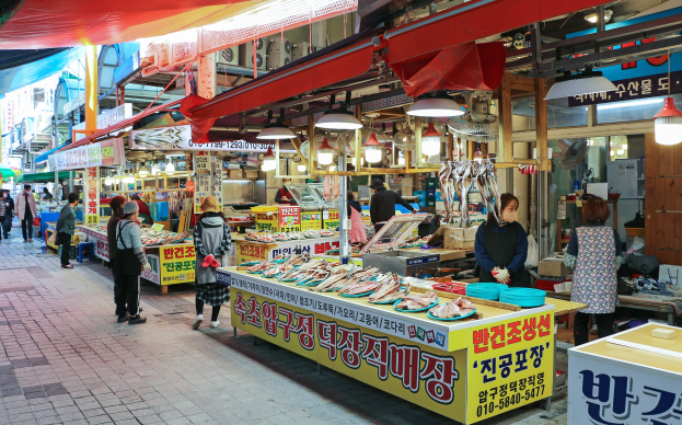 Ein belebter Straßenmarkt in Seoul, Südkorea, mit Menschen, die gehen, Tischen mit verschiedenen Gegenständen wie Lampen und Schildern und Gebäuden unter einem klaren blauen Himmel.