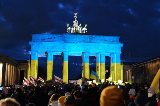 Eine Menschenmenge steht vor dem Brandenburger Tor in Berlin, Deutschland, mit Fahnen und Plakaten in den Händen, einem Banner auf der rechten Seite und dem Tor mit Statuen und Säulen geschmückt unter einem bewölkten Himmel.
