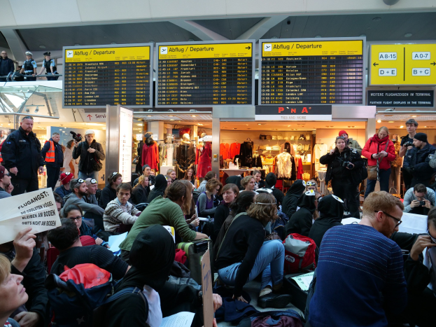 Eine große Gruppe von Menschen in einem Flughafen, einige sitzen mit Taschen und Papieren, andere stehen, mit Texttafeln und Schaufensterpuppen im Hintergrund, was auf eine Demonstration hindeutet.
