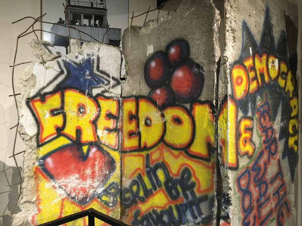 Eine Wand mit Graffiti, das in fetten schwarzen Buchstaben "Freiheit und Demokratie" lautet und unten ein Geländer sowie im Hintergrund ein Gebäude mit Fenstern zeigt.