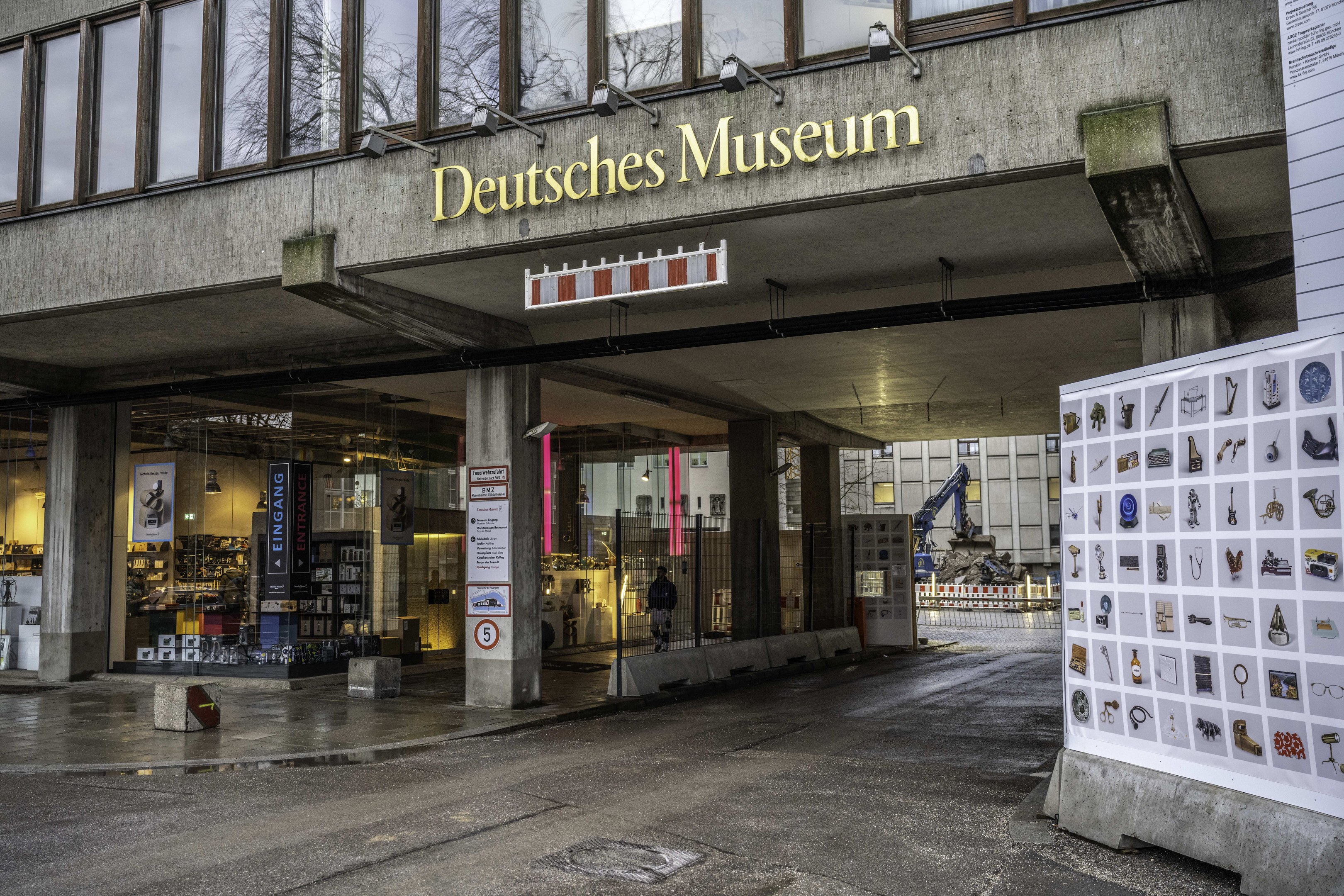 Großes Gebäude mit Glasfenstern und Säulen, beschriftet mit "Deutsches Museum" in Berlin, Deutschland, mit einer informativen Displaytafel auf der rechten Seite und anderen Gebäuden, Bäumen und Himmel im Hintergrund.