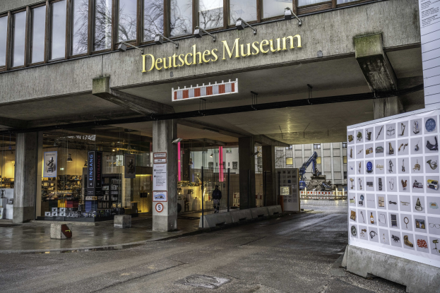 Großes Gebäude mit Glasfenstern und Säulen, beschriftet mit "Deutsches Museum" in Berlin, Deutschland, mit einer informativen Displaytafel auf der rechten Seite und anderen Gebäuden, Bäumen und Himmel im Hintergrund.