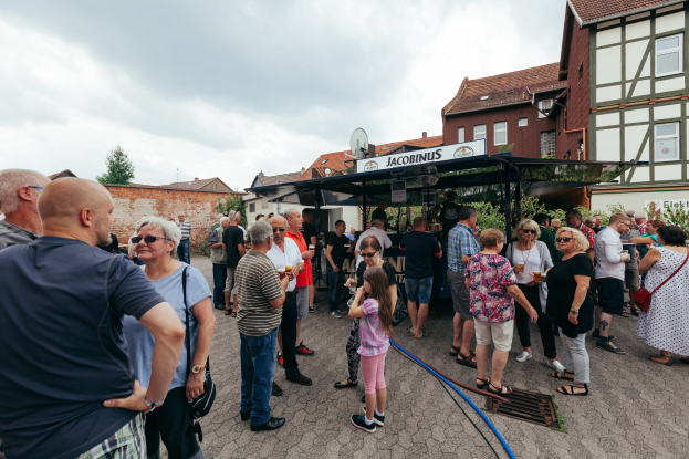 Gruppe von Menschen auf einem Outdoor-Bierfest, die vor einem Gebäude mit Fenstern, Bäumen und einem Schuppen mit einer Namensplakette im Hintergrund stehen.