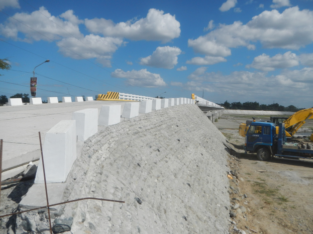 Baustelle mit einem großen Haufen Betonblöcke, einem Fahrzeug, einem Zaun, verstreuten Steinen, einer Baumgruppe, einem Straßenschild, einer Stromleitung und einem bewölkten Himmel.