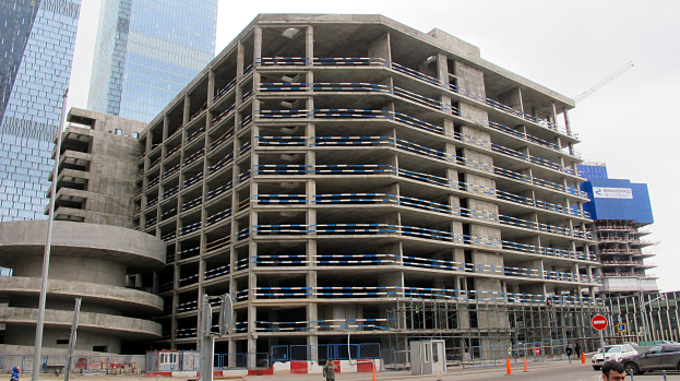 Ein großes Gebäude im Bau in einer städtischen Umgebung, umgeben von Wolkenkratzern, mit Fahrzeugen, Fußgängern, Verkehrsinfrastruktur und Baumaschinen unter einem klaren Himmel.