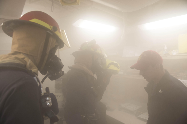Feuerwehrleute mit Helmen und Rauch, der aus ihren Mündern kommt, einer hält eine Kamera, während einer Trainingsrunde mit Hintergrundbeleuchtung und -ausrüstung.