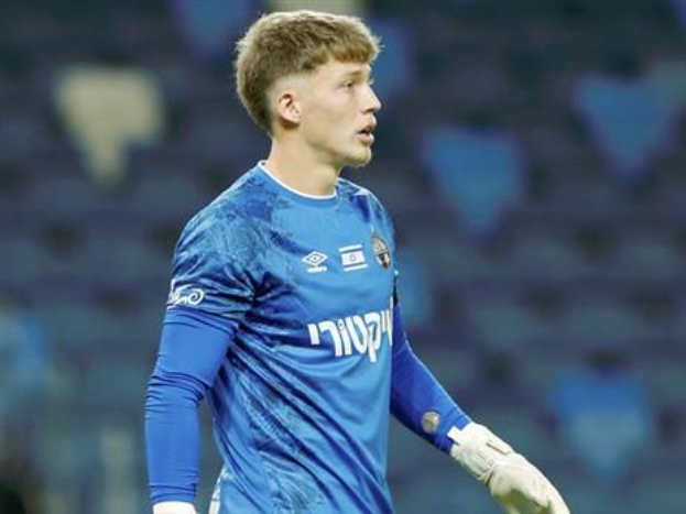 Ein junger Mann in einem blauen Torwarttrikot und weißen Handschuhen steht auf einem Fußballfeld, mit einem unscharfen Hintergrund, scheinbar mitten in einem Spiel.