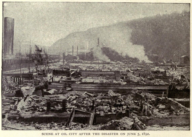 Schwarzes und weißes Foto einer Stadt nach einer Katastrophe am 5. Juni 1892, das Gebäude, Pfähle, Rauch und einen Hügel im Hintergrund zeigt, mit Text am unteren Rand.