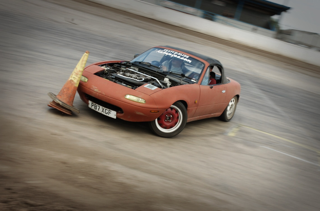 Eine Person in einem roten Auto auf einer Rennstrecke, die sich einem Verkehrskegel nähert, mit einem unscharfen Hintergrund, der hohe Geschwindigkeit andeutet.