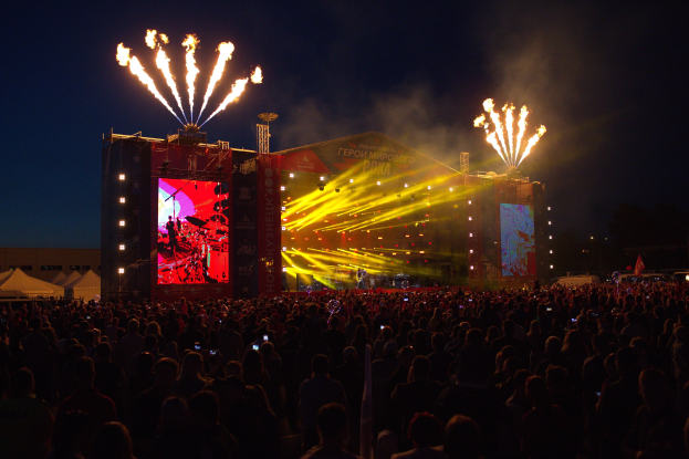 Eine große Menschenmenge auf einem Musikfestival mit beleuchteter Bühne und Performern, umgeben von Bannern, Zelten links und Feuerwerk am dunklen Himmel dahinter.
