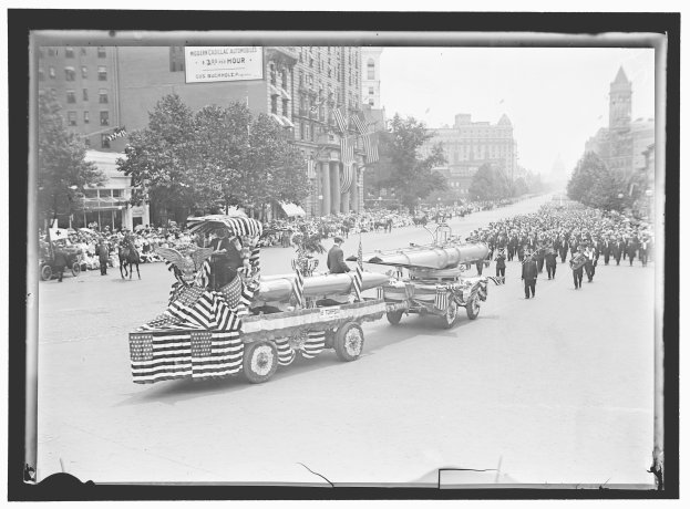 Schwarzes und weißes Foto eines Umzugs mit einem Feuerwehrauto in der Mitte, umgeben von Menschen mit Fahnen, sowie Bäumen, Gebäuden und einer Tafel mit Text im Hintergrund.