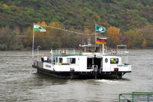 Eine Fähre mit Flaggen und Masten fährt einen Fluss hinunter, mit einem Geländer auf der rechten Seite und einem Hügel mit Bäumen im Hintergrund, und trägt einige Menschen an Bord.