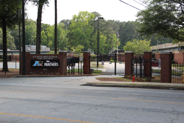 Schuleingang mit einem Schild, umgeben von Bäumen, Straßenmöbeln, Fahrzeugen und Gebäuden unter einem klaren blauen Himmel.