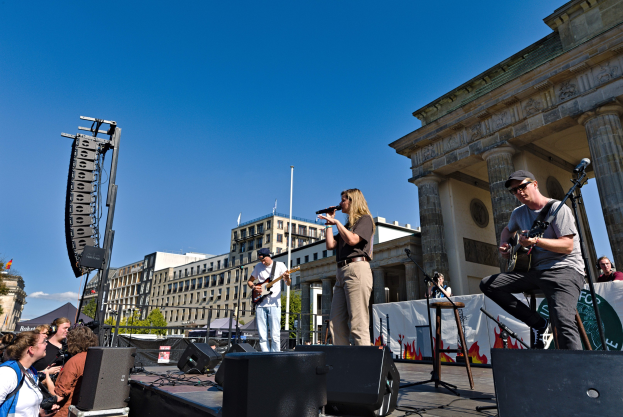 Eine Gruppe von Menschen, die auf einer Bühne Musik spielen, mit dem Brandenburger Tor im Hintergrund, begleitet von Lautsprechern und Equipment unter einem klaren Himmel.