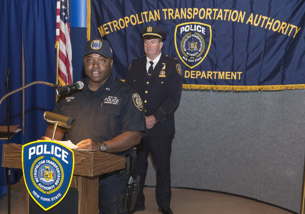 Zwei Polizisten stehen an einem Podium mit einem Mikrofon, einer hält Papiere; ein Banner, eine Flagge und ein Tisch sind im Hintergrund sichtbar.