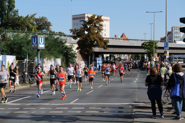 Gruppe von Menschen, die bei einem Marathon auf einer baumgesäumten Straße laufen, mit Laternenpfählen, Schildern, Fahrrädern, einem Zaun, Gras, einer Brücke, Gebäuden und einem klaren blauen Himmel.