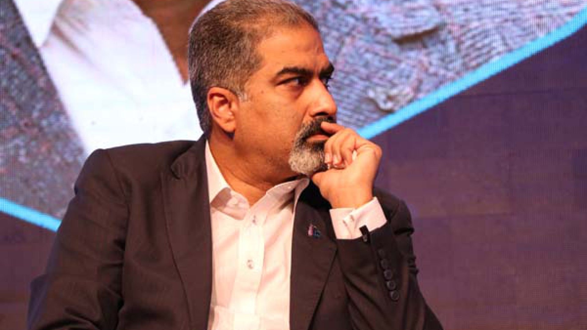 Ein Mann im Anzug sitzt auf einer Bühne und denkt nach, vor einem Bildschirm.