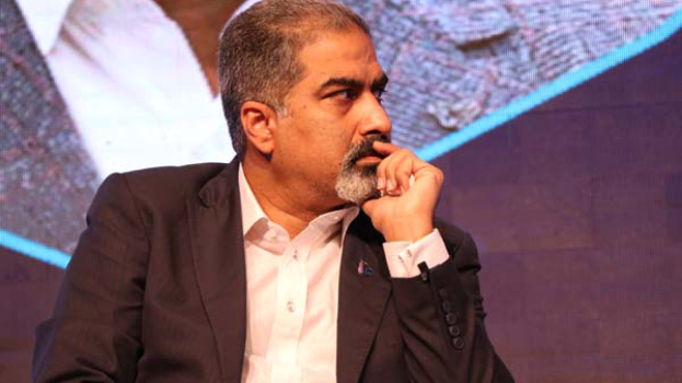 Ein Mann im Anzug sitzt auf einer Bühne und denkt nach, vor einem Bildschirm.