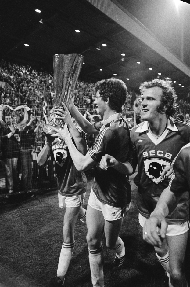 Schwarzes und weißes Bild von Fußballspielern, die mit einem Pokal auf einem Feld feiern, umgeben von einer Menge hinter einem Zaun unter Stadionlichtern.