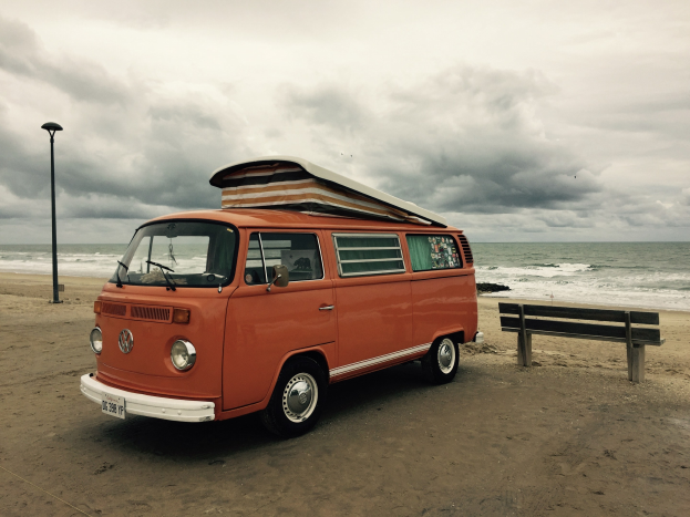 Ein oranger VW-Bus auf einem Strand in der N├Ąhe des Meeres geparkt, mit einer Bank und einem Laternenmast in der N├Ąhe, unter einem bew├Âlktem Himmel mit dem Wasser im Hintergrund.