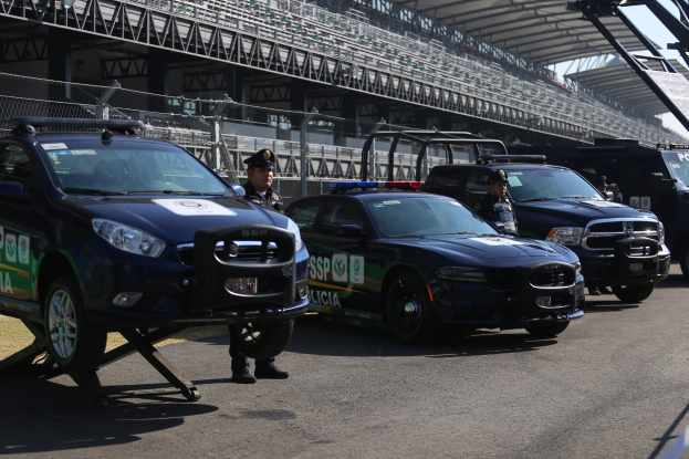 Polizeiautos vor einer Rennstrecke geparkt, uniformierte Personen auf der Straße stehend, Stadion und Absperrung im Hintergrund.