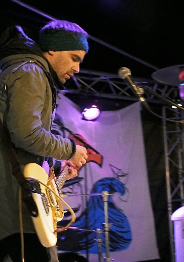 Ein Mann in einer Jacke und einer Mütze spielt eine E-Gitarre auf der Bühne, mit einem Mikrofon vor ihm, Scheinwerfern und einem Banner im Hintergrund und einem Schlagzeug zu seiner Rechten.