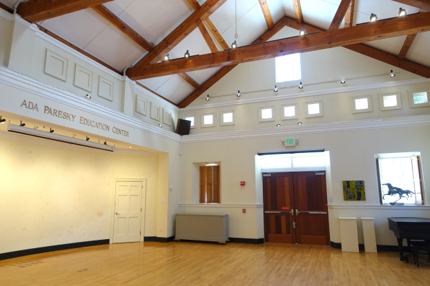 Ein geräumiger Raum mit Holzfußboden und weißen Wänden, der ein Piano und verschiedene Gegenstände enthält, identifiziert als Ada Paresky Education Center.