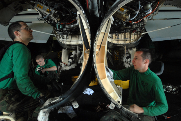 Zwei Männer in grünen T-Shirts arbeiten an einem Jet-Triebwerk in einer Halle, umgeben von verschiedenen Ausrüstungsstücken.