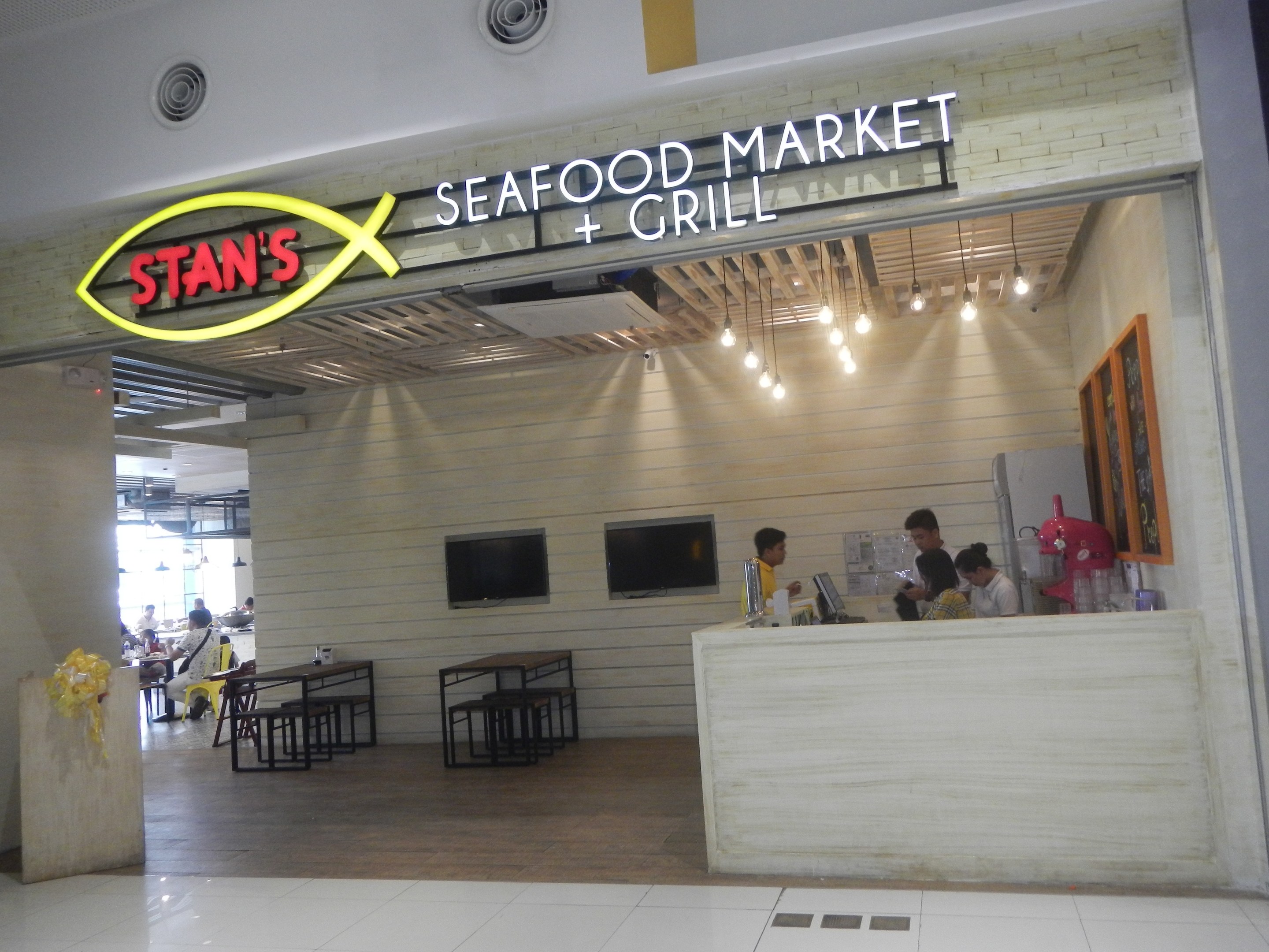 Innenansicht von Stans Seafood Market & Grill mit sitzenden und stehenden Gästen, einem Tisch mit Gegenständen darauf auf der rechten Seite, einem gerahmten Foto an der Wand, an der Wand montierten Fernsehern und Deckenbeleuchtung.