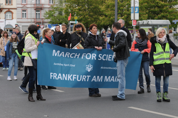 Eine bunte Gruppe von Menschen marschiert auf einer Straße, hält ein "March for Science Frankfurt am Main"-Schild hoch, mit Bäumen, Gebäuden und einem klaren Himmel im Hintergrund.