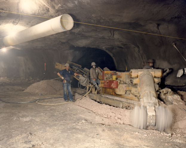 Gruppe von Männern in Helmen bei der Arbeit in einem dunklen Stollensystem mit Maschinen und Rohren.