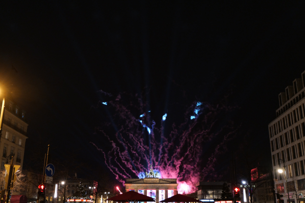 Eine belebte Straßenkreuzung in Berlin, Deutschland, mit dem Brandenburger Tor im Hintergrund, beleuchtet von Gebäudelichtern, Straßenlaternen und Feuerwerk am Nachthimmel.