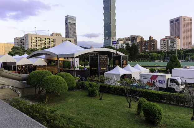Eine Gruppe von Zelten in einem Park umgeben von Grünanlagen, mit Menschen, Fahrzeugen, Gebäuden und einem klaren Himmel im Hintergrund, wahrscheinlich aufgenommen auf dem Hong Kong International Film Festival.