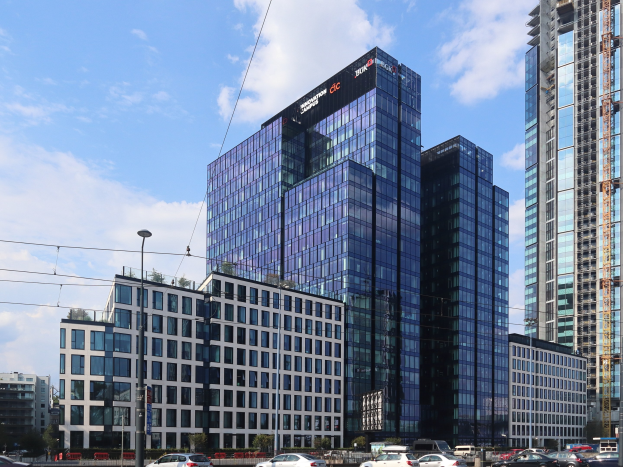 Großes Gebäude mit vielen Fenstern, als Deutsche Bank Hauptsitz identifiziert, umgeben von Stadtinfrastruktur einschließlich Straßenmasten, Lampen, Fahrzeugen, Grünflächen und einem bewölkten Himmel.