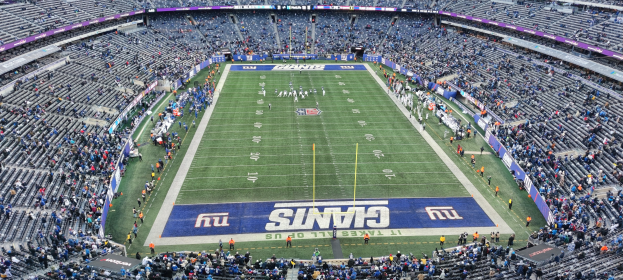 Ein voller Fußballstadion mit Spielern auf dem Feld, Torpfosten und Texttafeln, umgeben von leeren Sitzplätzen.