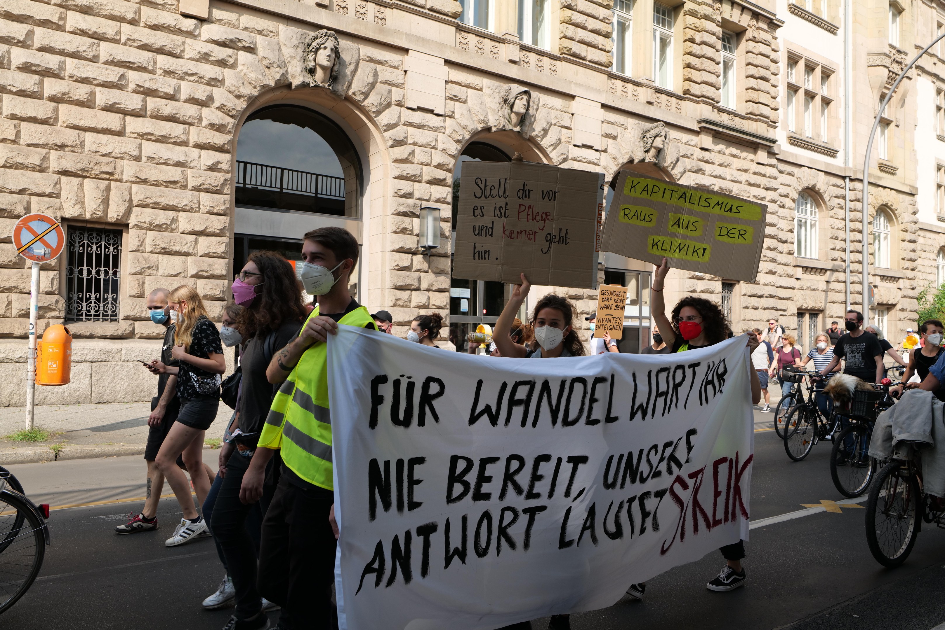 Eine Gruppe von Menschen marschiert auf einer Straße bei einer Demonstration, einige halten Schilder und andere fahren Fahrräder, im Hintergrund ein historisches Gebäude mit Rundbogenfenstern, Säulen und Skulpturen.