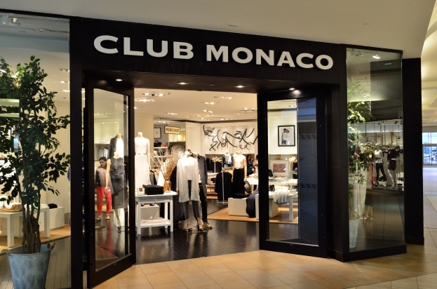 Ein Club Monaco-Laden in einem Einkaufszentrum mit Glaswänden und -türen, Schaufensterpuppen in Kleidern, Tischen mit Waren, einem Topf mit einer Pflanze und Deckenleuchten.
