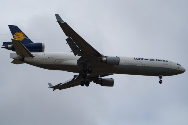 Lufthansa Cargo Boeing 777-300ER im Flug mit sichtbarem Text auf dem Rumpf, ausgefahrene Tragflächen, aktive Triebwerke, vor einem Himmel mit weißen Wolken.
