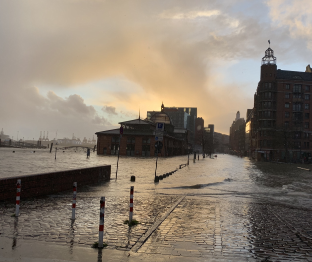 Überschwemmte Straße in Hamburg, Deutschland, mit Wasser, das die Straße, Pfosten, Schilder, Gebäude, eine Brücke und einen bewölkten Himmel bedeckt.