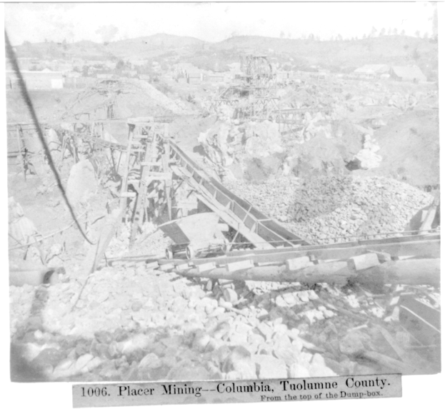 Ein Schwarz-Weiß-Foto eines Kohlebergwerks in Kolumbien mit ein paar Gebäuden, Bäumen und Bergen im Hintergrund und Text am unteren Bildrand.