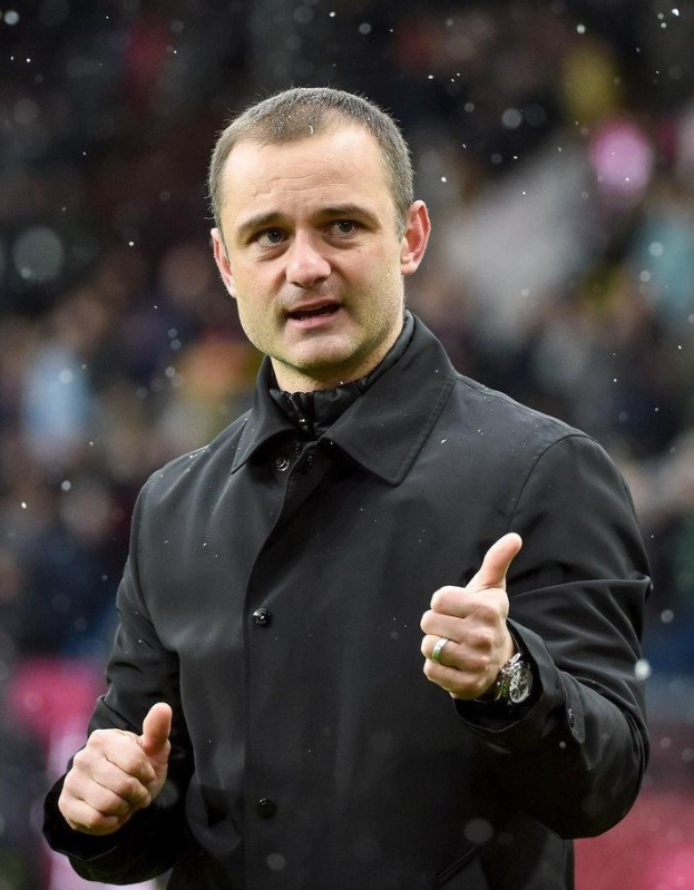 Ein Mann in einer schwarzen Jacke zeigt den Daumen hoch, trägt eine Uhr und hat einen unscharfen Hintergrund, was darauf hindeutet, dass er ein Fußballtrainer ist.