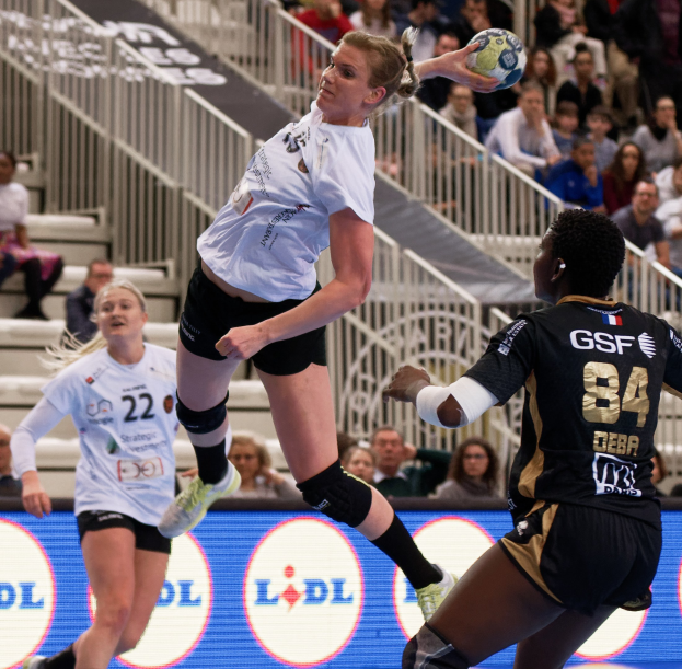 Eine Gruppe von Frauen, die Handball auf einem Platz spielt, mit einem Ball in der Luft, Zuschauern auf der Treppe im Hintergrund und einer "Handball-Weltmeisterschaft 2019 - Tag 2"-Plakette.