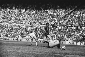 Schwarze-Weiß-Fotografie von Fußballspielern im Einsatz auf einem Feld mit Zuschauern im Hintergrund und einer Bundesliga-1978-79-Ajax-1-1-Hannover-96-Banner.