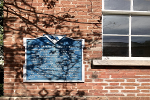 Ein altes Bankgebäude mit einer blauen Plakette an der Seite, einem Fenster auf der rechten Seite und einem Baum auf der linken Seite.