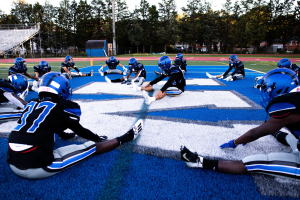 Gruppe junger Männer in Sportkleidung, Helmen und Schuhen sitzend auf einem Fußballfeld mit Bäumen, Pfosten, Gebäuden und einem klaren blauen Himmel im Hintergrund, Treppe mit Geländern auf der linken Seite.