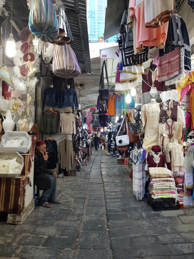 Ein belebter Straßenmarkt in der Altstadt von Jerusalem mit Ständen, die Kleidung, Taschen, Lampen und andere Artikel verkaufen, während Menschen im Hintergrund browsen und interagieren.