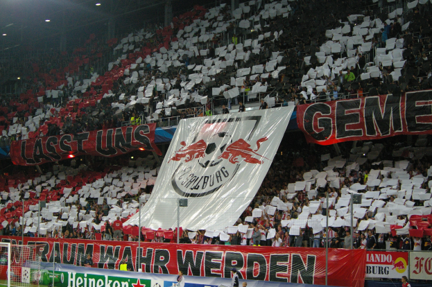 Fußballstadion mit Sitzplätzen und Stehplätzen, Banner mit Text und Logos, ein Tor und Überdachungsbeleuchtung.