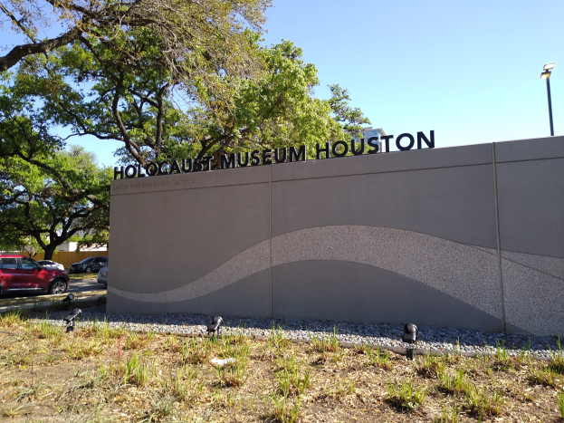 Außenansicht des Houston Holocaust Museums mit einer Gedenkwand, Steinen, Landschaftsgestaltung, Bäumen, Straßenverkehr, einem Straßenschild und einem bewölkten Himmel.