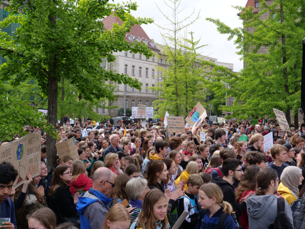 Große Menschenmenge protestiert vor einem Gebäude in Berlin, hält Schilder, mit Bäumen, Fahrzeugen, einem Lautsprecher und Himmel im Hintergrund.